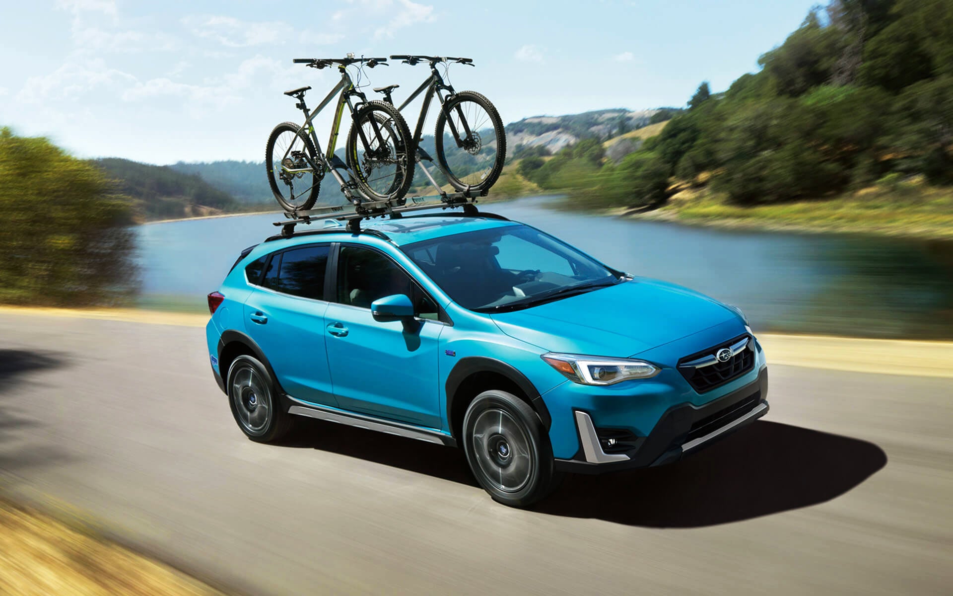 A blue Crosstrek Hybrid with two bicycles on its roof rack driving beside a river | Fuccillo Subaru in Watertown NY